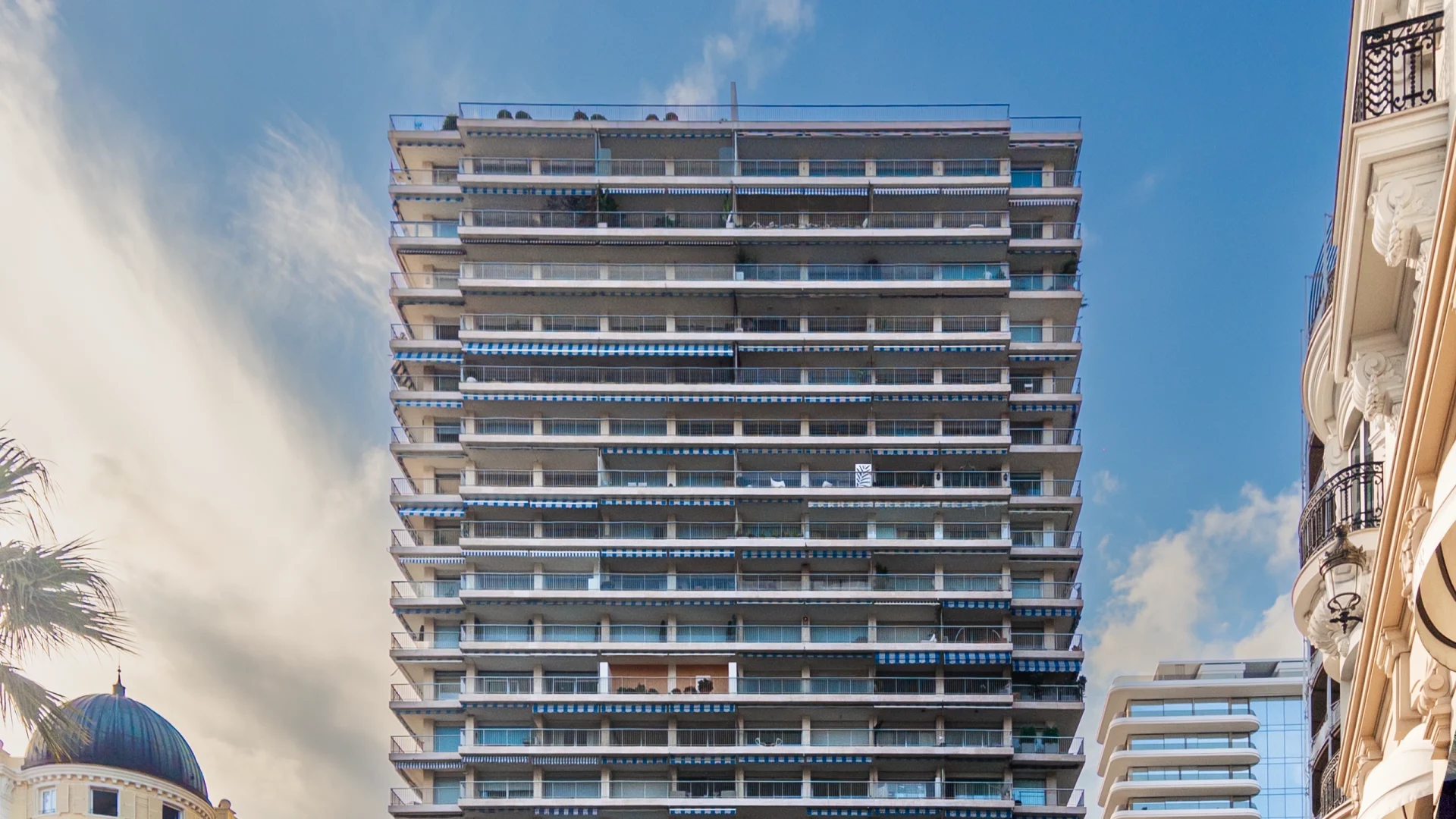 A photo of the sun tower residence in Monaco from street level. In this photo you see the residence in the middle, it is a white building, in the background you see the 26 Carré d'Or residence, Hotel de Paris, Palais de la Scala, and Hermitage hotel