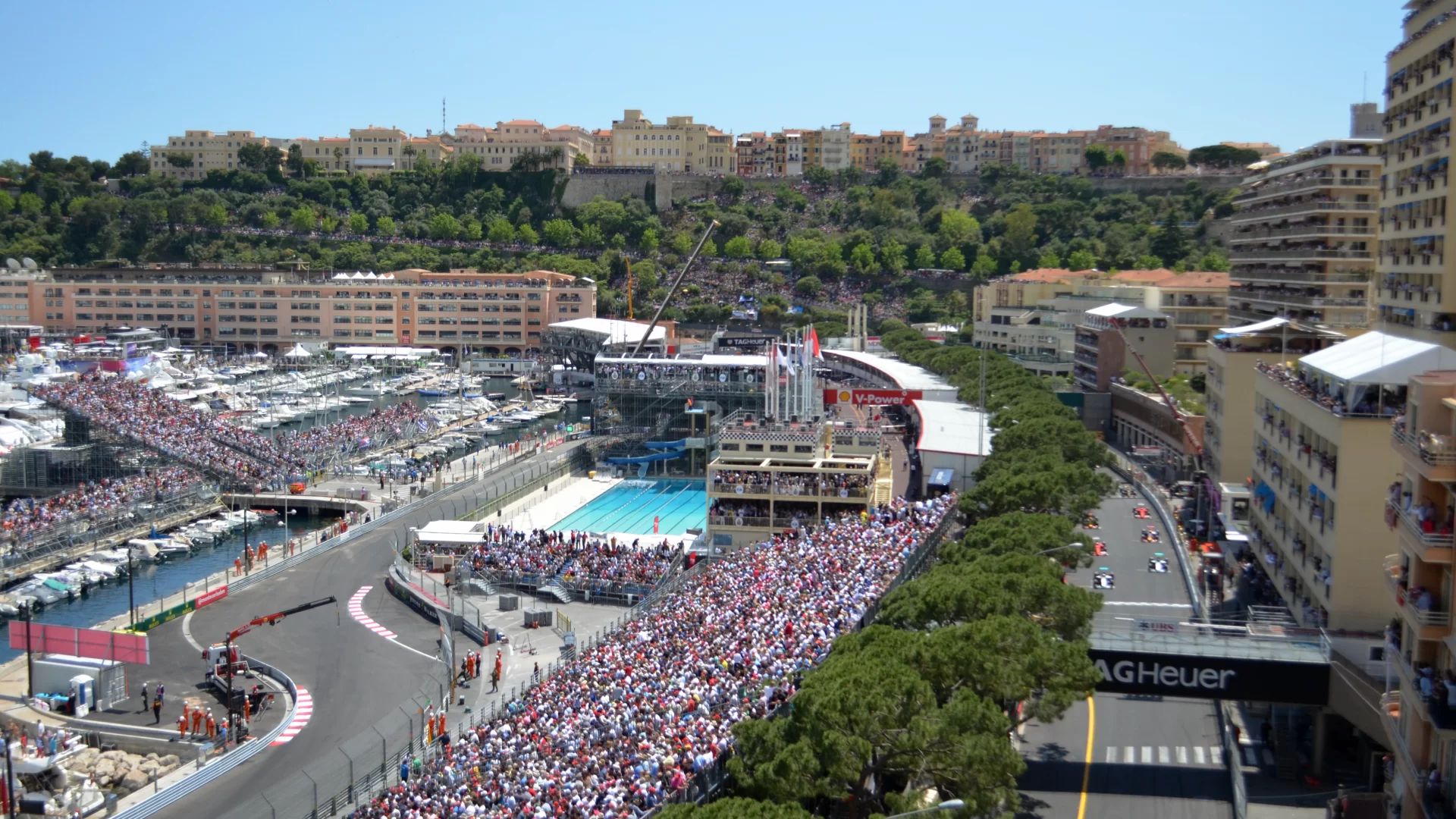 This is the view from Ermanno Palace during formula 1 in Monaco. You see many people in the stands surrounding the track, Le Roche, A pool, and surrounding buildings.