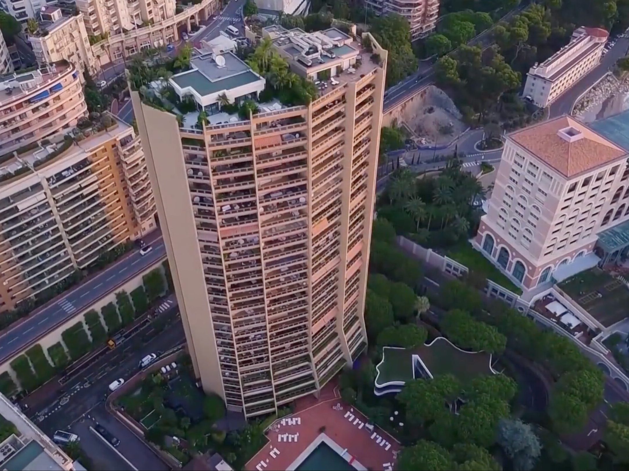 A photo of the Roccabella Residence in Monaco. In this Photo you see the residence, a pool, greenery, and surrounding streets and buildings such as the Monte Carlo Bay Hotel