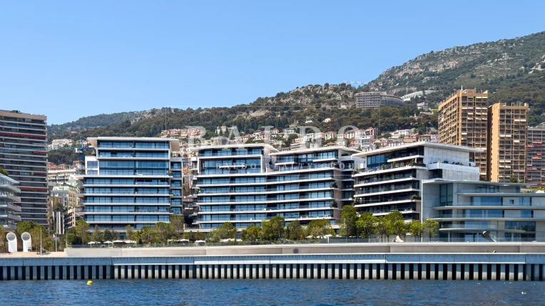 A front ocean view of the residence building Les Jardins d'Eau. You have a close view of the Mareterra district in Monaco with the residence, gardens, walkway, and ocean in full view