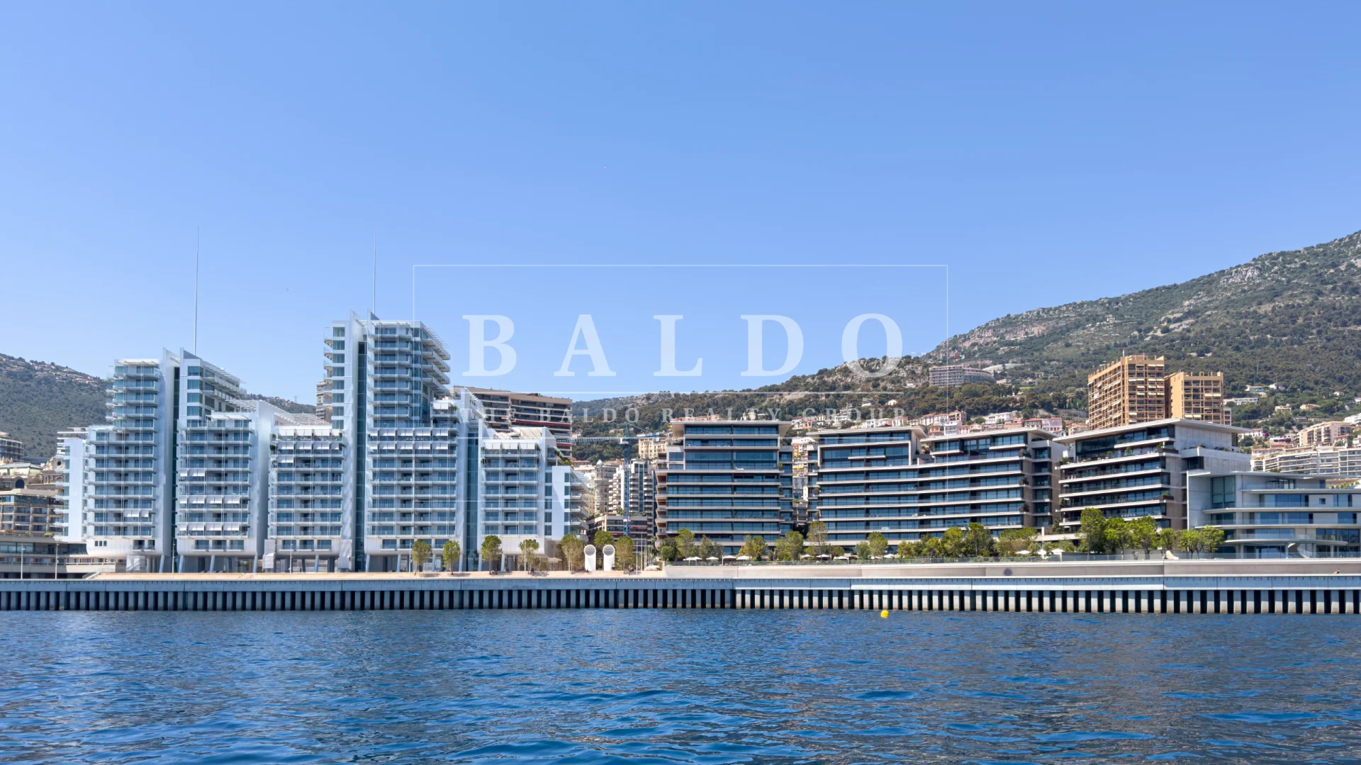 Le Renzo and Les Jardins d'Eau residence in Mareterra Monaco. In this photo you see the two residences, Le Renzo on the right, Les Jardins d'Eau on the left. In front of the residences are the Mediterranean Sea, Esplanade et Promenade Prince Jacques walkway, and land reclamation needed for the project. The sky is blue and in the background you have mountains.