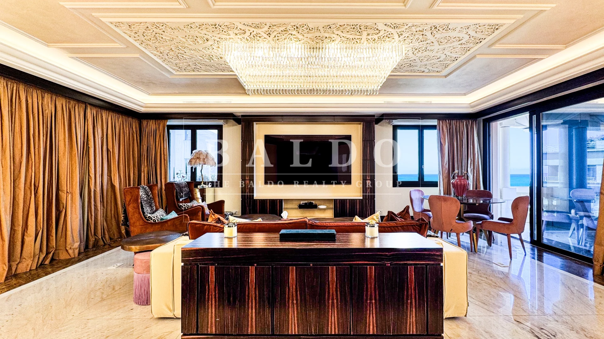 A living room in the Floristan residence. In this photo you see a wide living room with a cabinet and couch in the forefront. Along the rear of the living room you see a Tv with a window on each side with sea views. At the top you see a large crystal chandelier and in the far right you have a table and door to the balcony.