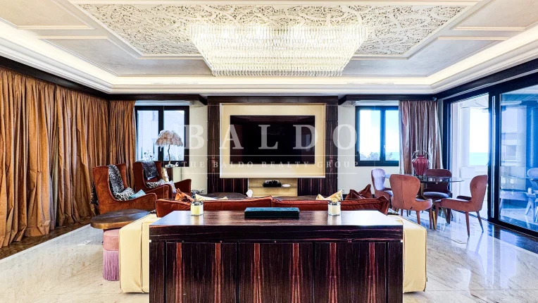 A living room in the Floristan residence. In this photo you see a wide living room with a cabinet and couch in the forefront. Along the rear of the living room you see a Tv with a window on each side with sea views. At the top you see a large crystal chandelier and in the far right you have a table and door to the balcony.