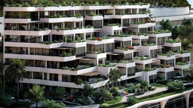 A photo of the L'Exotique residence located in Jardin Exotique, Monaco. In the photo you see the residence which is white in a blocky style, surrounded by greenery with a mountain and street behind it.