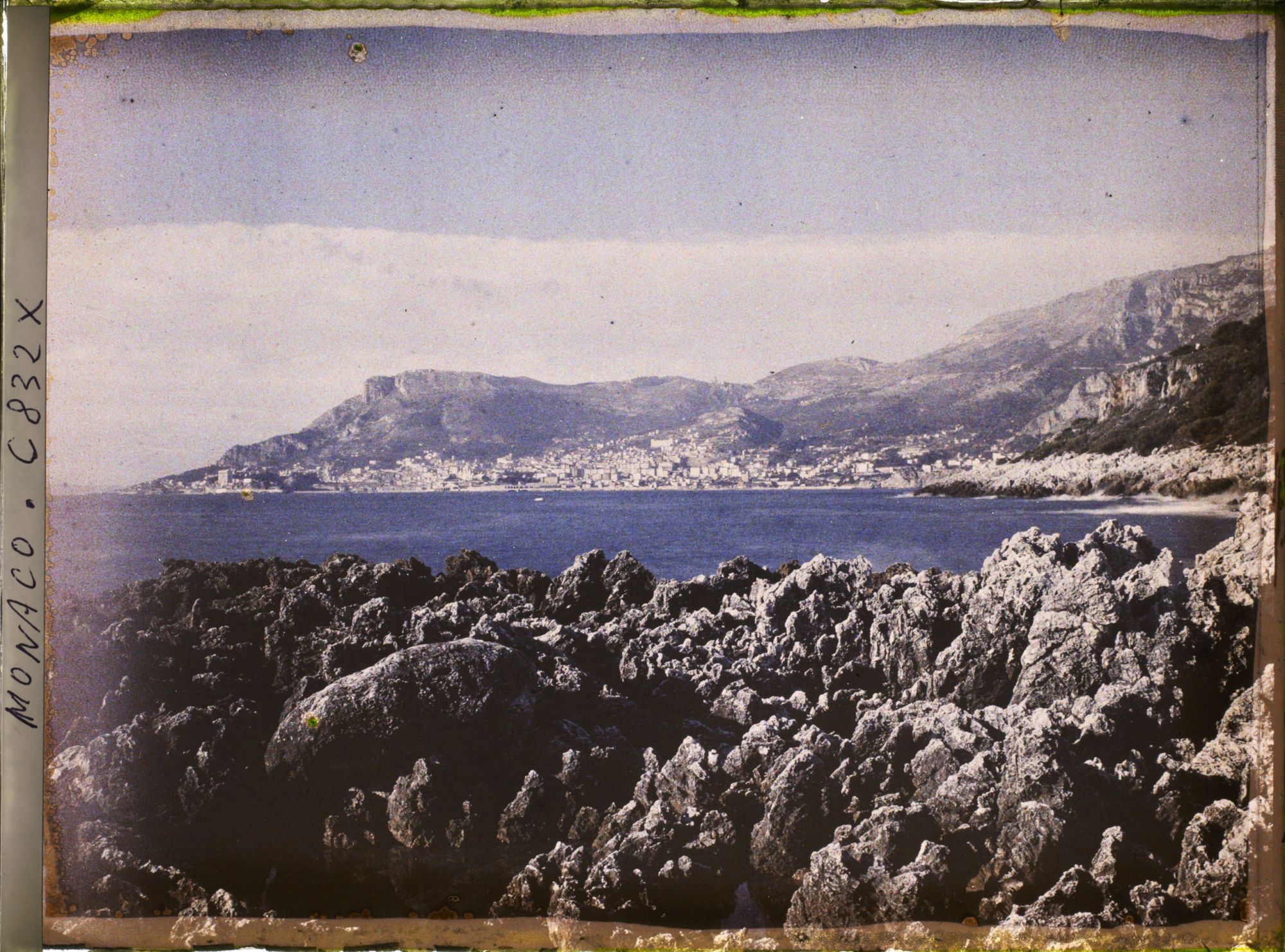 Rocky shoreline of Cap Martin with Monaco across the bay, early colour photograph
